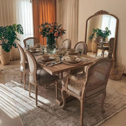 غرفة طعام كلاسيكية تضم كراسي بظهر خيزران وطاولة خشبية مستطيلة مع مرآة كبيرة ونباتات منزلية في أجواء مشمسة.