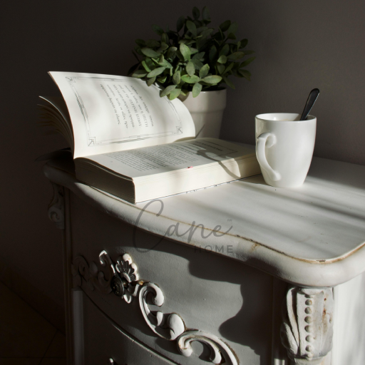 كومودينو كلاسيكي أبيض بتفاصيل حفر يدوية يضم كتاباً مفتوحاً وفنجان قهوة تحت إضاءة طبيعية دافئة.