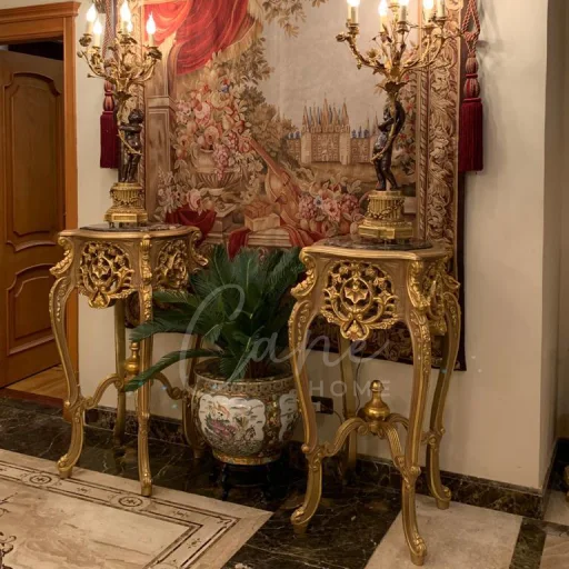 طاولات جانبية تحف مذهبة بتصميم أعمدة ملكية تحمل شمعدانات ذهبية أمام جدارية كلاسيكية فاخرة.